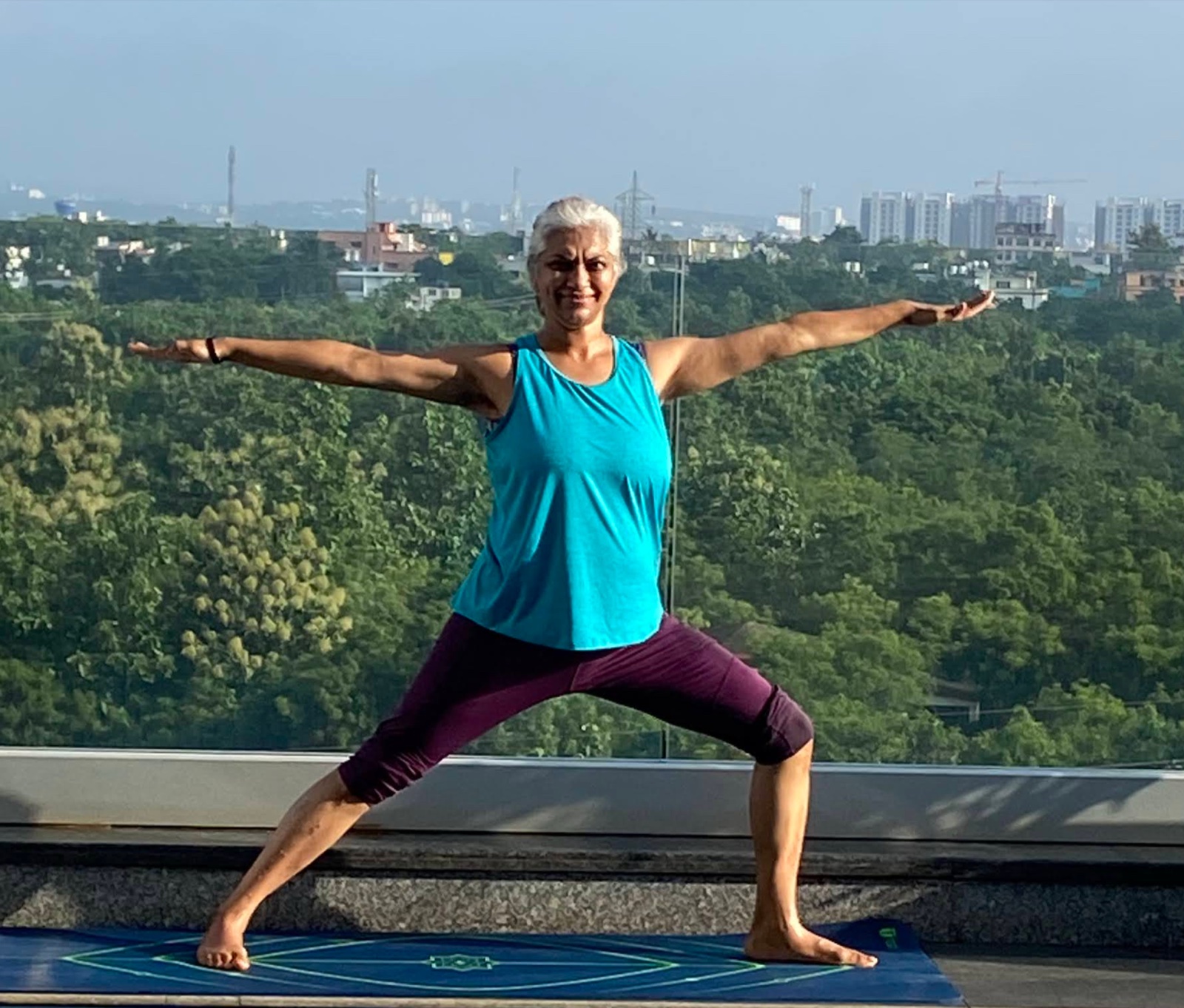 Sowmya Ayyar practising the Warrior II yoga pose on a rooftop terrace, treetops and city skyline behind her